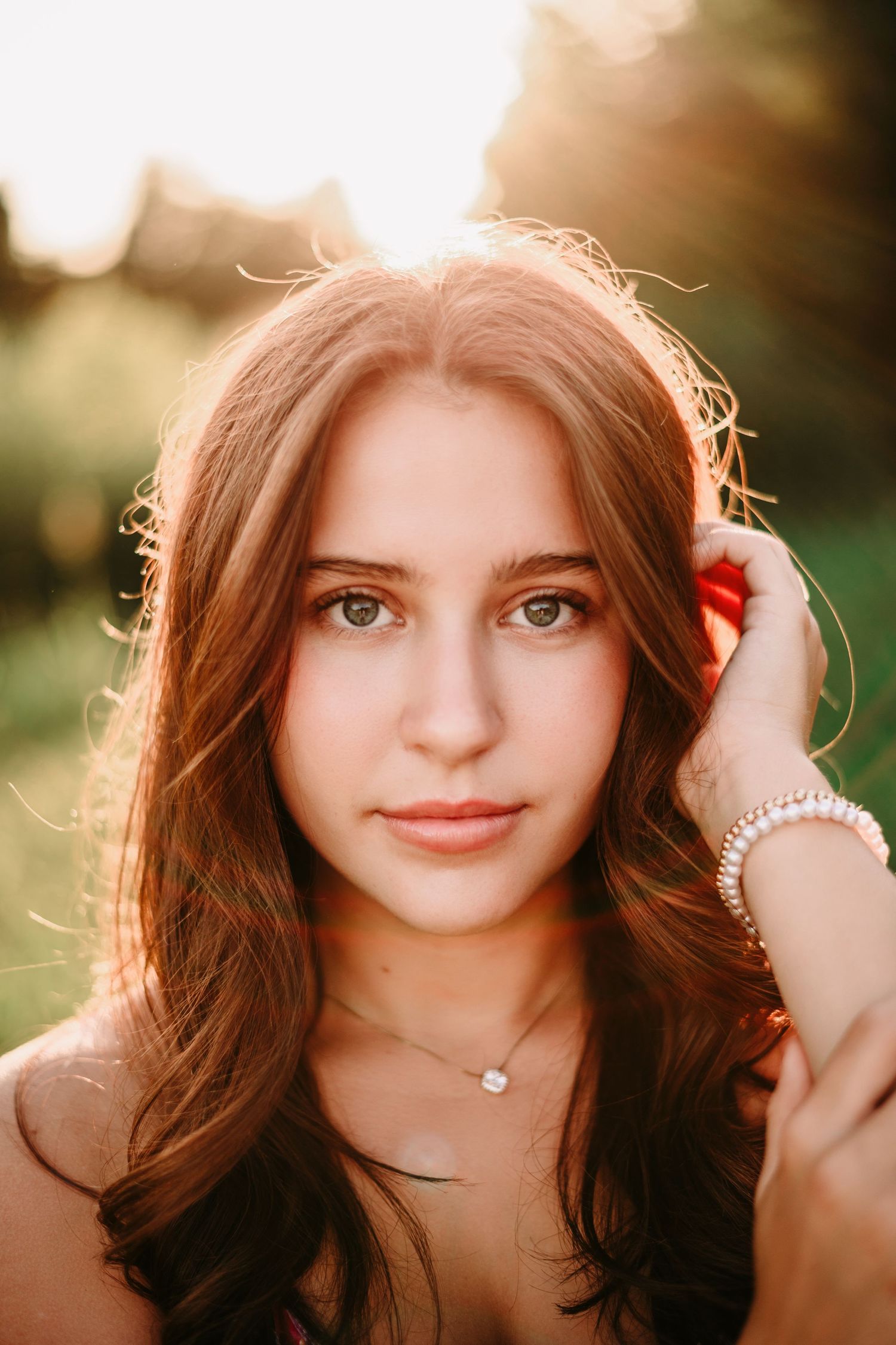 Young woman with brown wavy hair and green eyes poses outdoors in golden sunset light wearing a pearl bracelet.