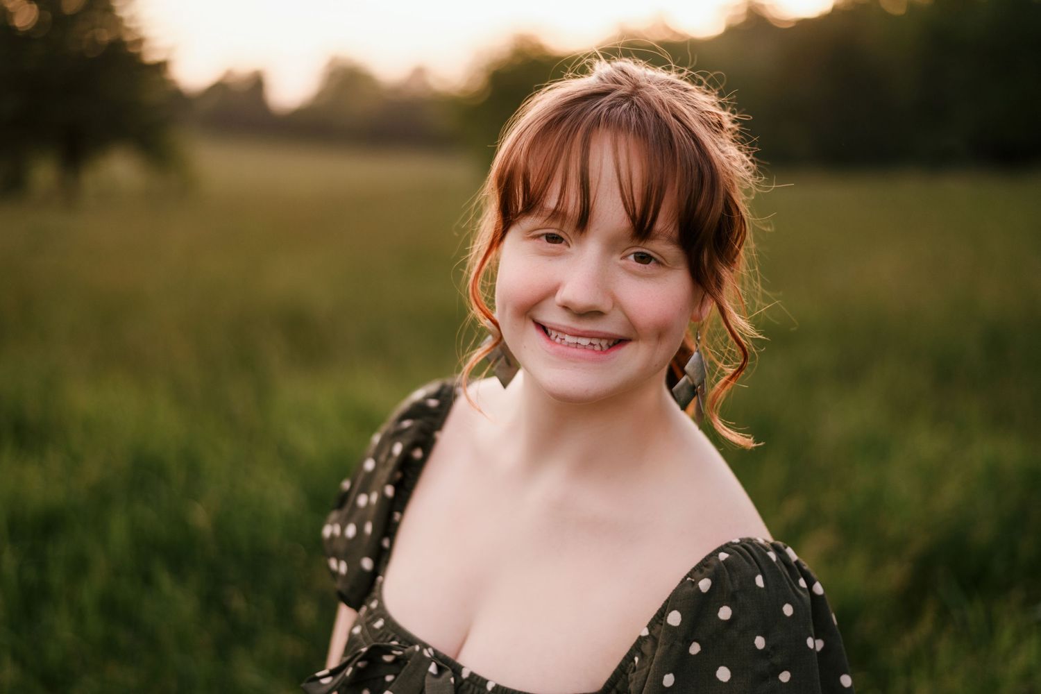Smiling young woman with bangs in polka dot dress poses in a sunlit green field at golden hour.
