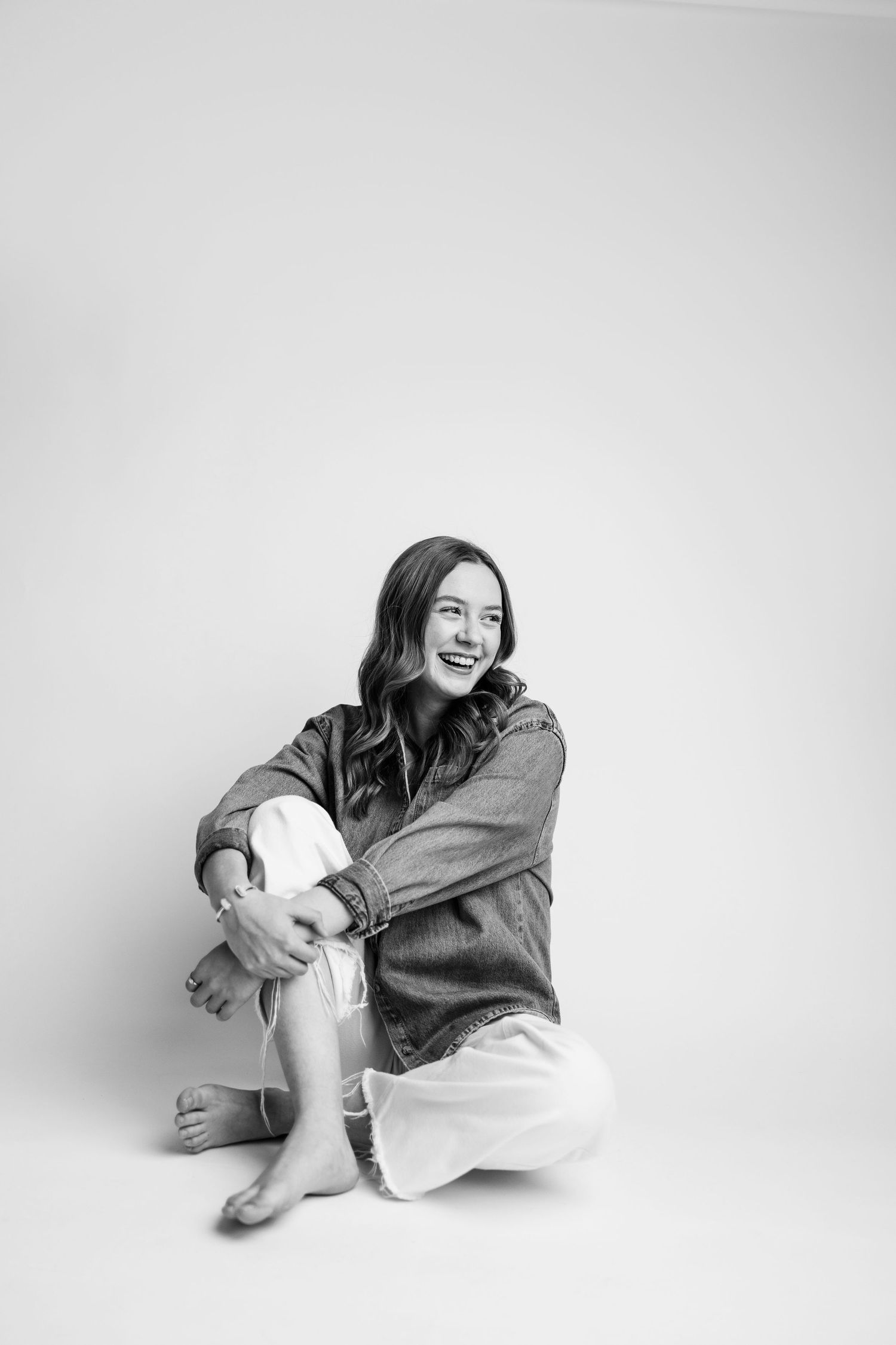 Black and white photo of a smiling young woman sitting cross-legged on the floor wearing a denim jacket and white pants.