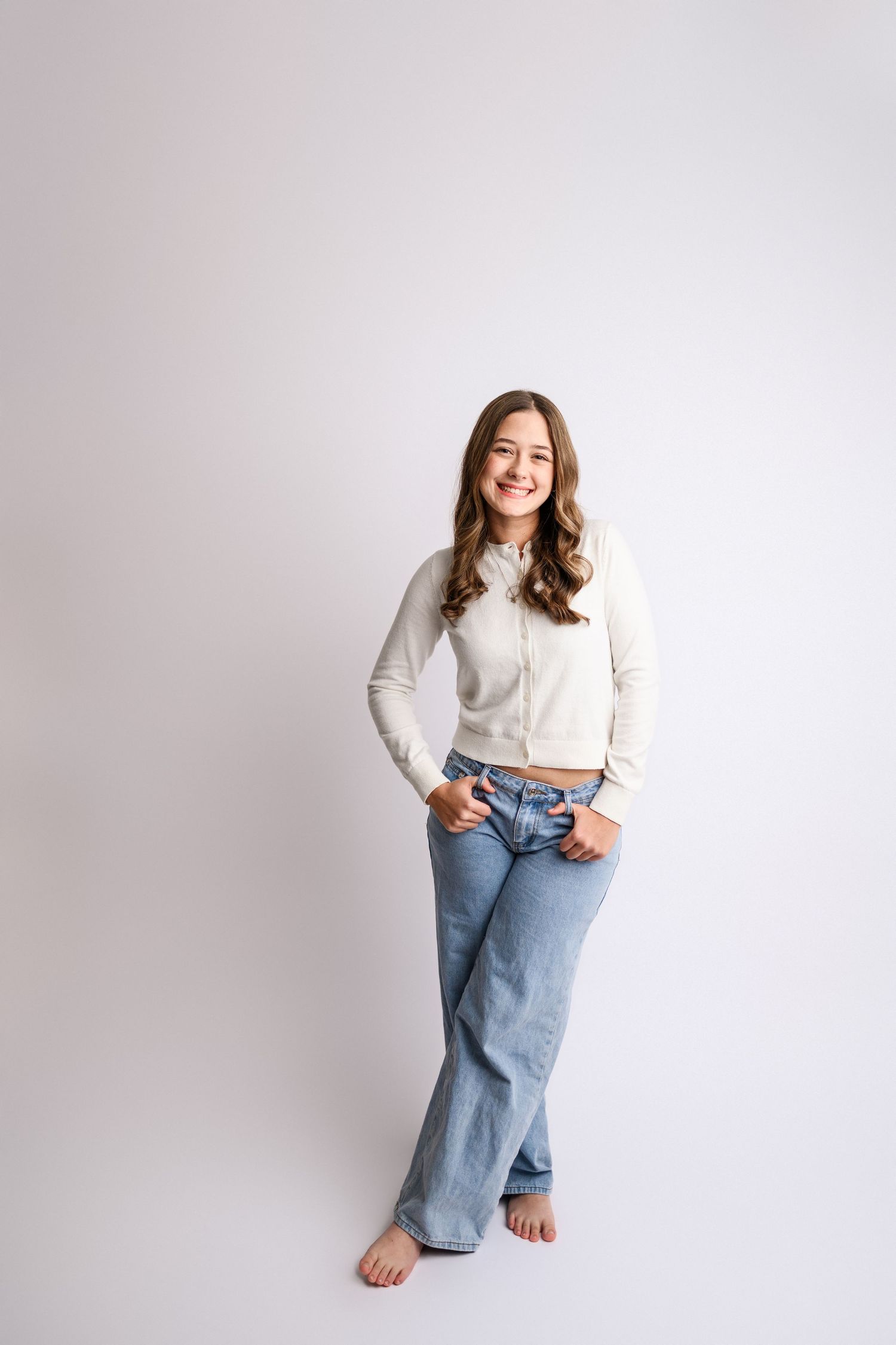 Smiling young woman in white cardigan and blue jeans posing barefoot against a white studio background.