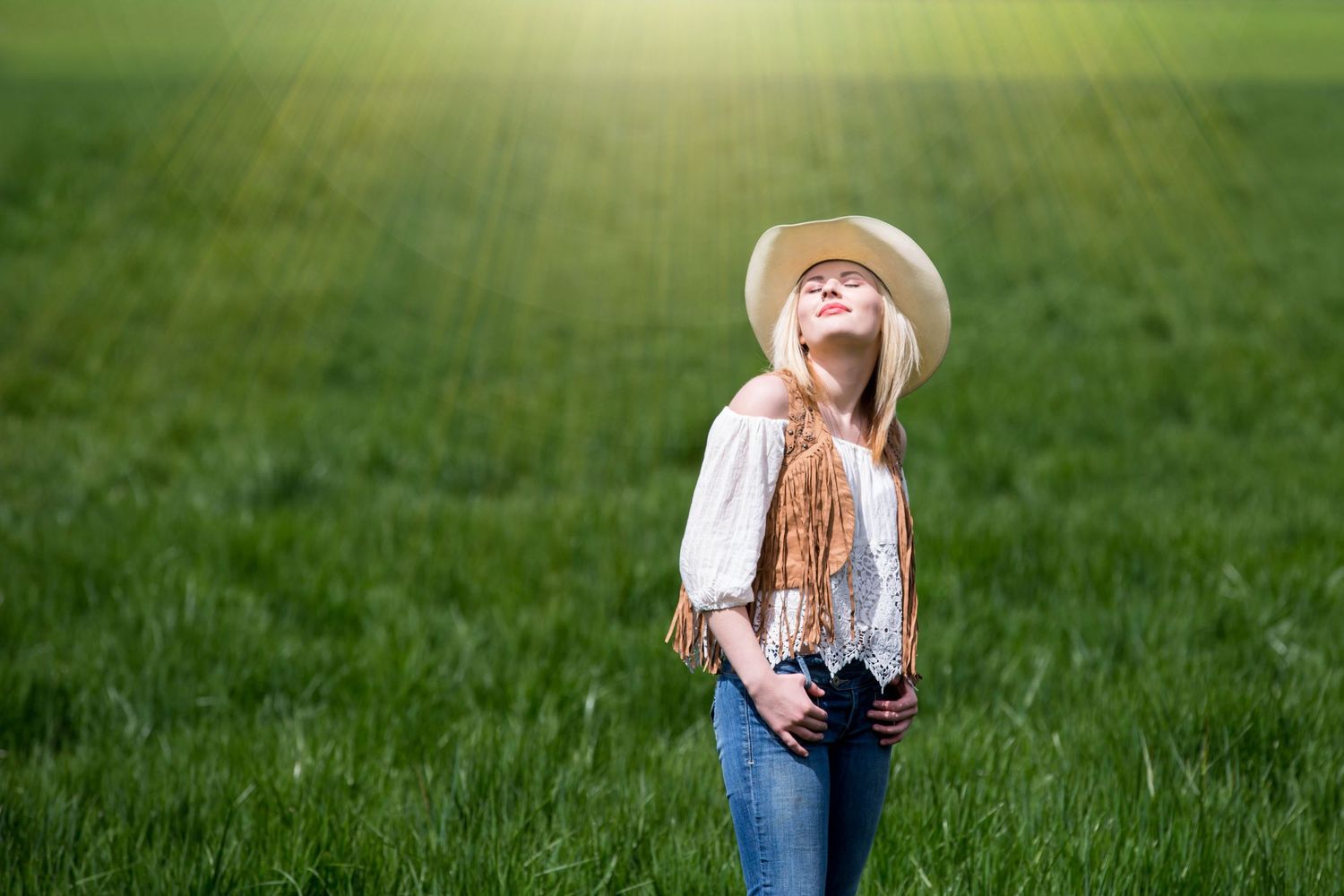 Person in cowgirl hat and jeans standing in sunlit green field.