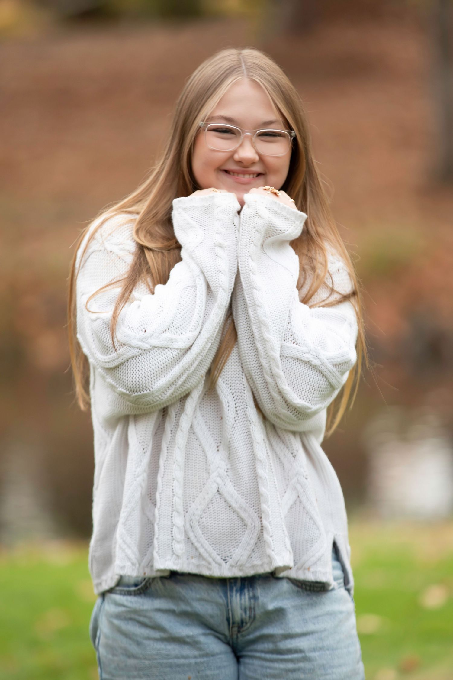 Smiling girl with long hair and glasses wearing a white cable-knit sweater and jeans, posing outdoors with autumn foliage behind her.