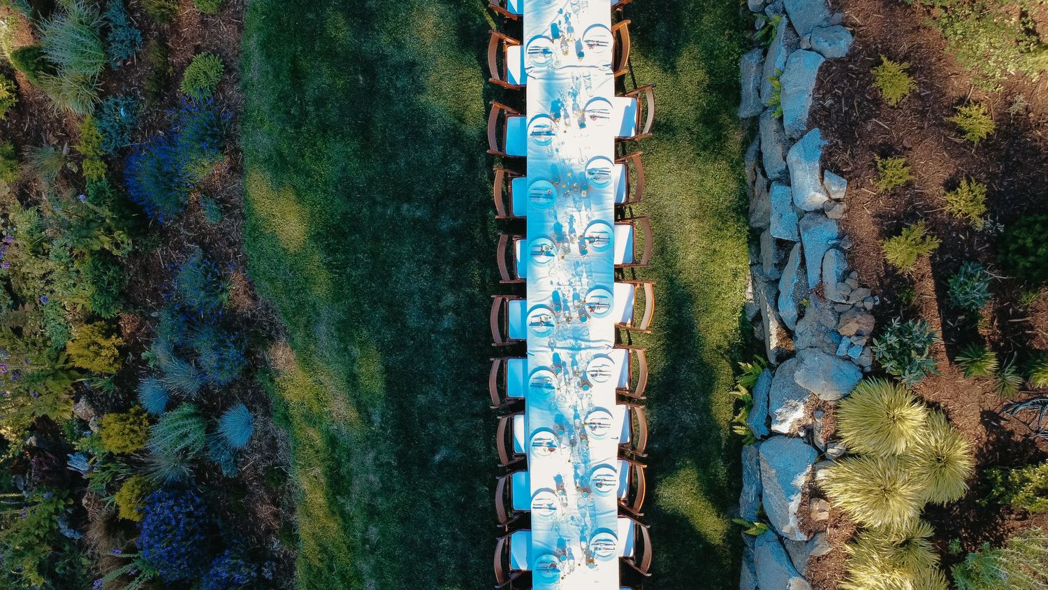 Aerial drone view of a long elegantly set outdoor dining table surrounded by lush green gardens.