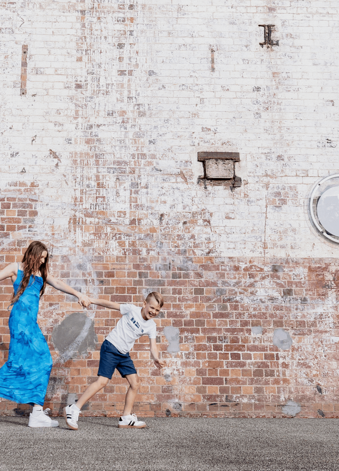 A woman in a blue dress and a boy in a white tee hold hands, playing near a rustic brick wall outdoors.