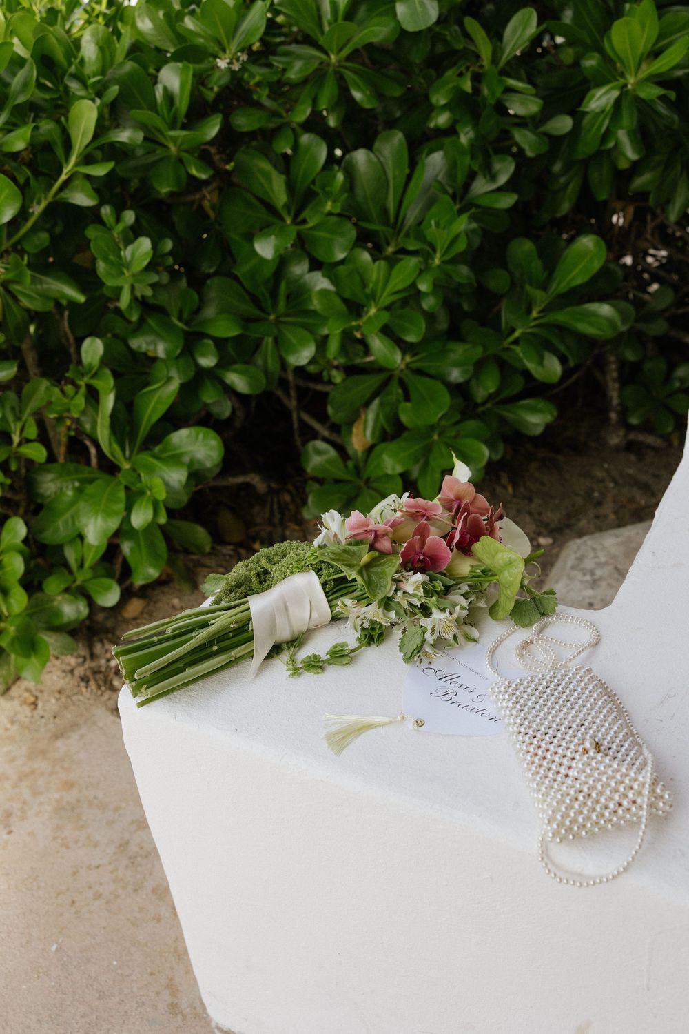 Bridal bouquet with pink and white flowers, pearl clutch, and wedding card on white surface with lush green foliage backdrop.