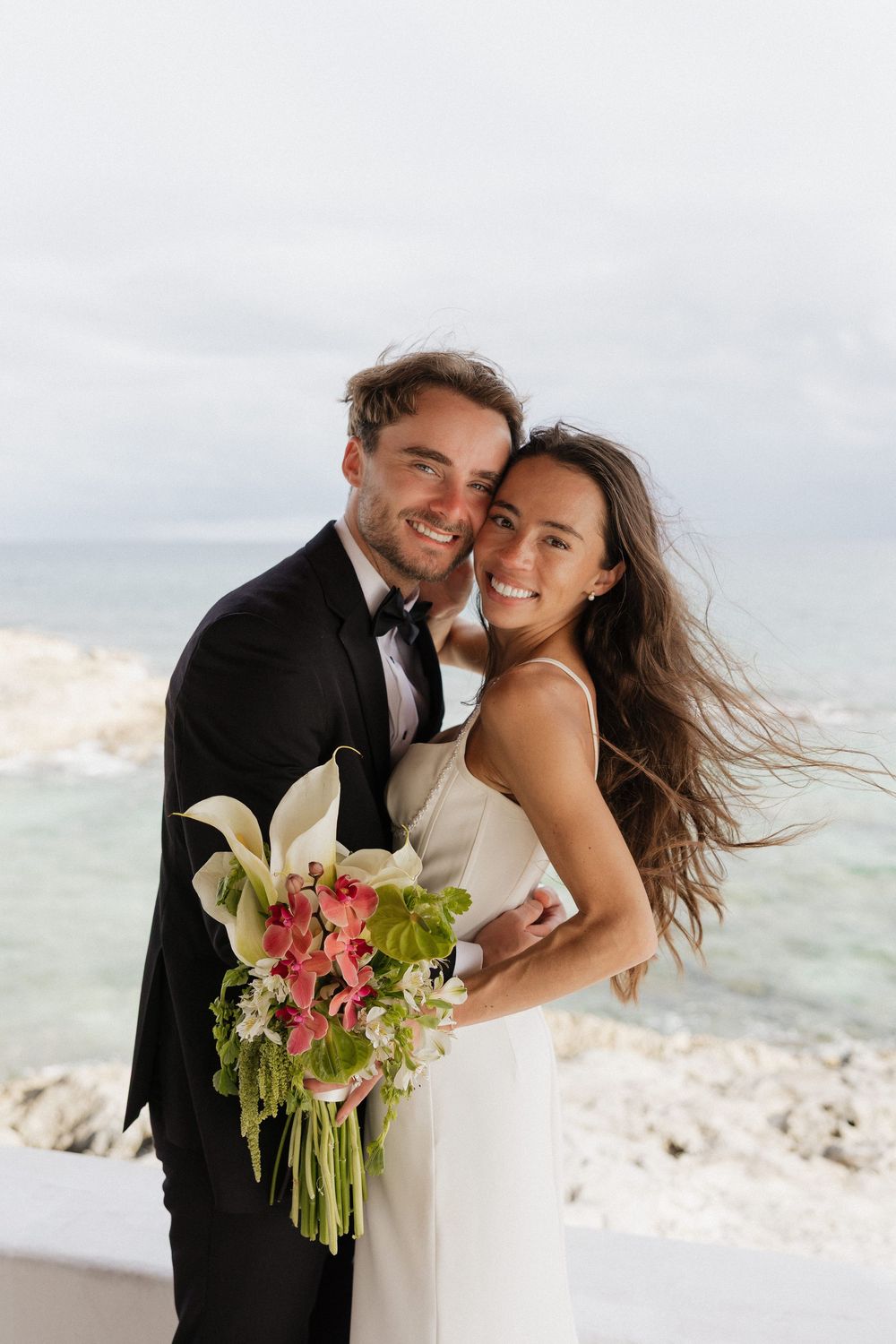 Smiling bride and groom embrace by rocky ocean shore, she holds tropical bouquet, he wears black tuxedo, white dress.