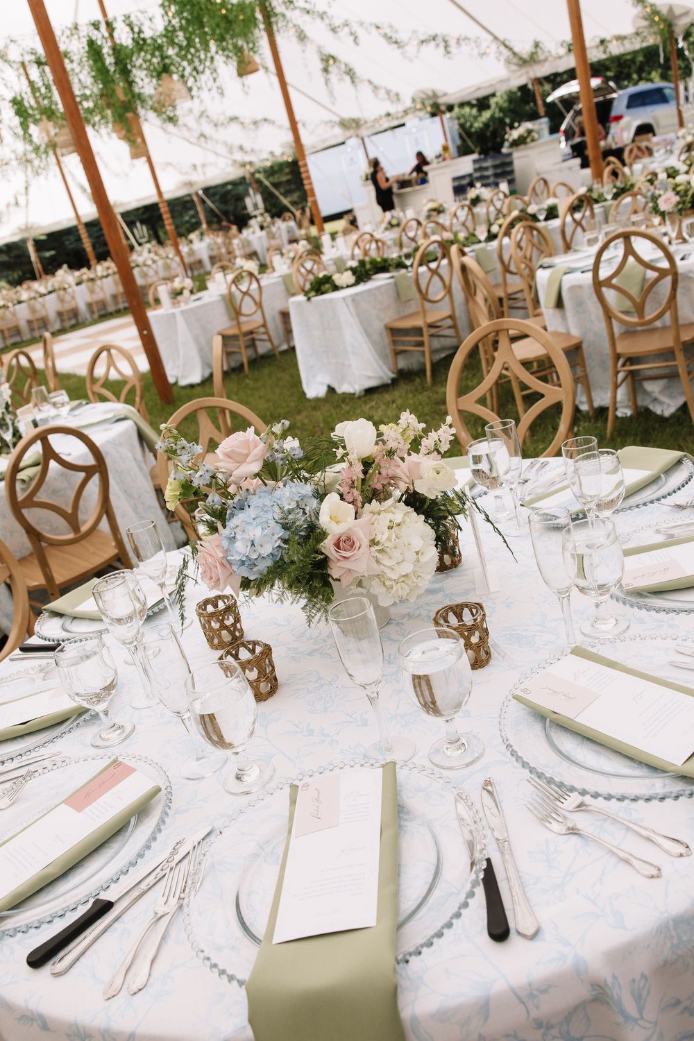 Elegant wedding reception tables with floral centerpieces, gold chairs, and sage green napkins under a white tent.