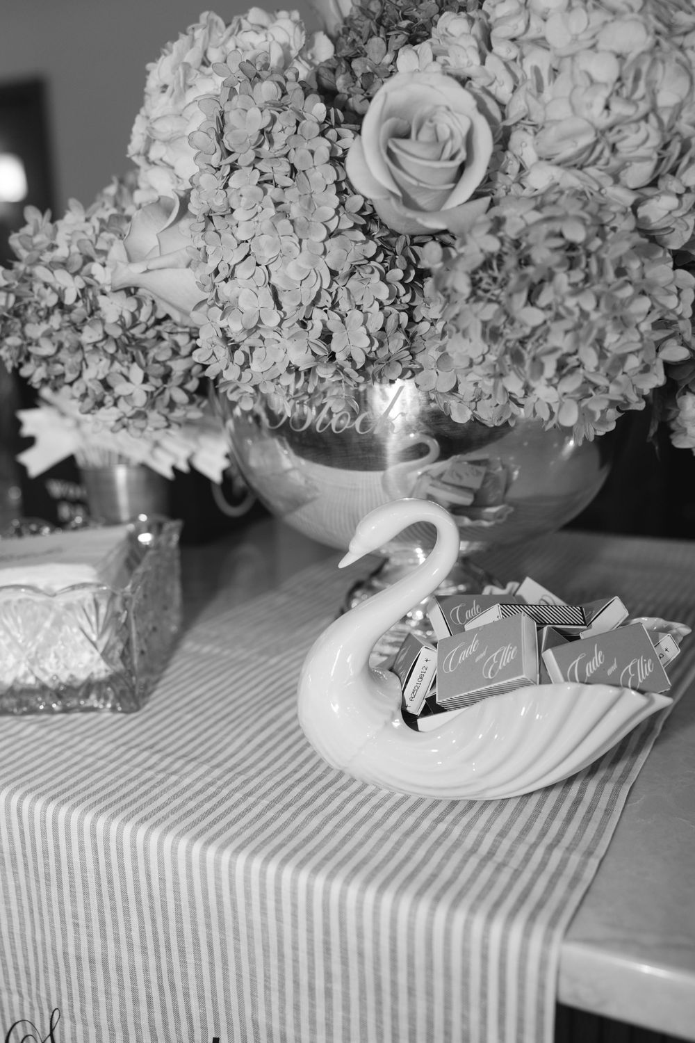Black and white photo of floral centerpiece with roses and hydrangeas in a vase beside a swan dish with chocolates.