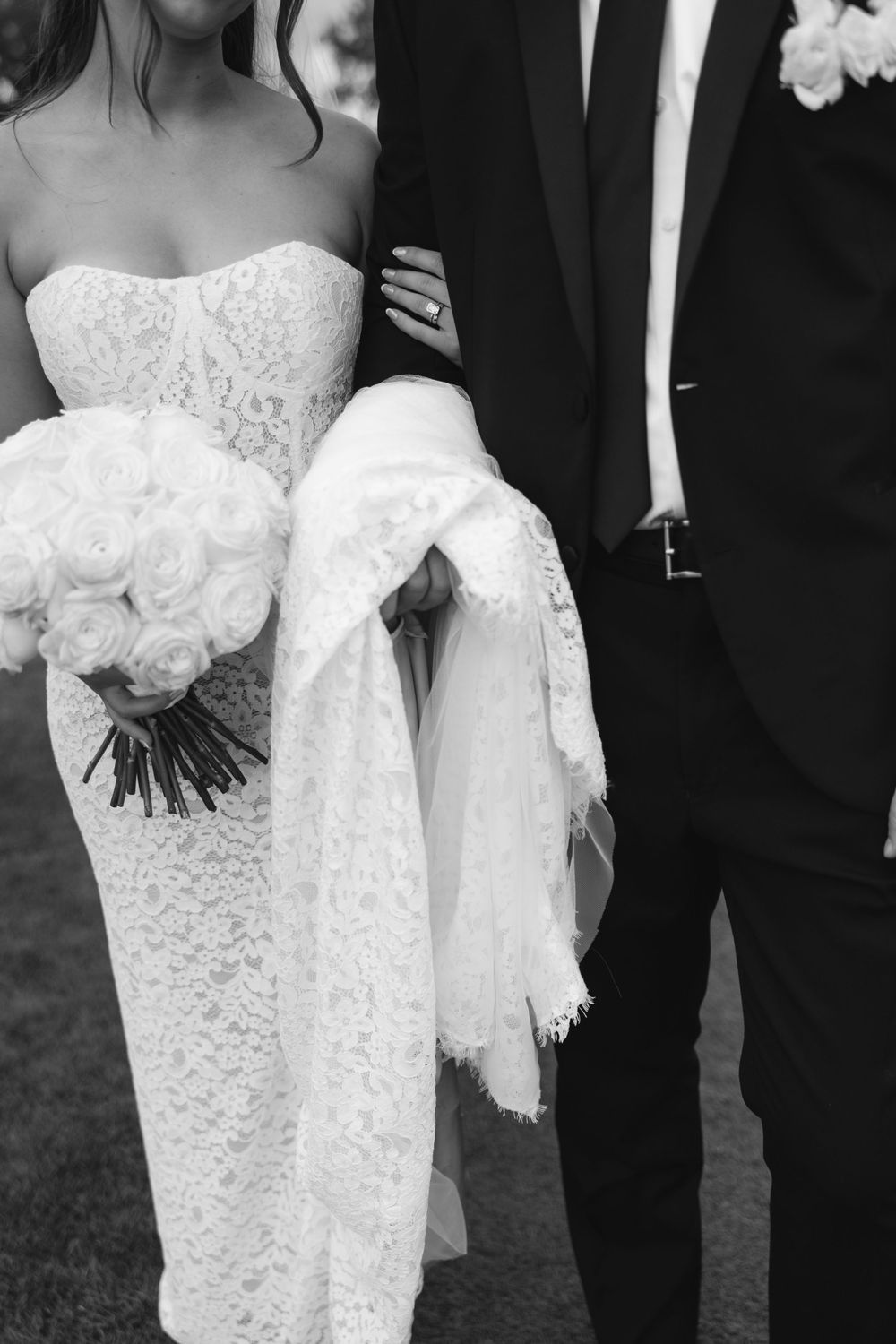 Black and white photo of bride in lace gown holding rose bouquet standing beside groom in dark suit.