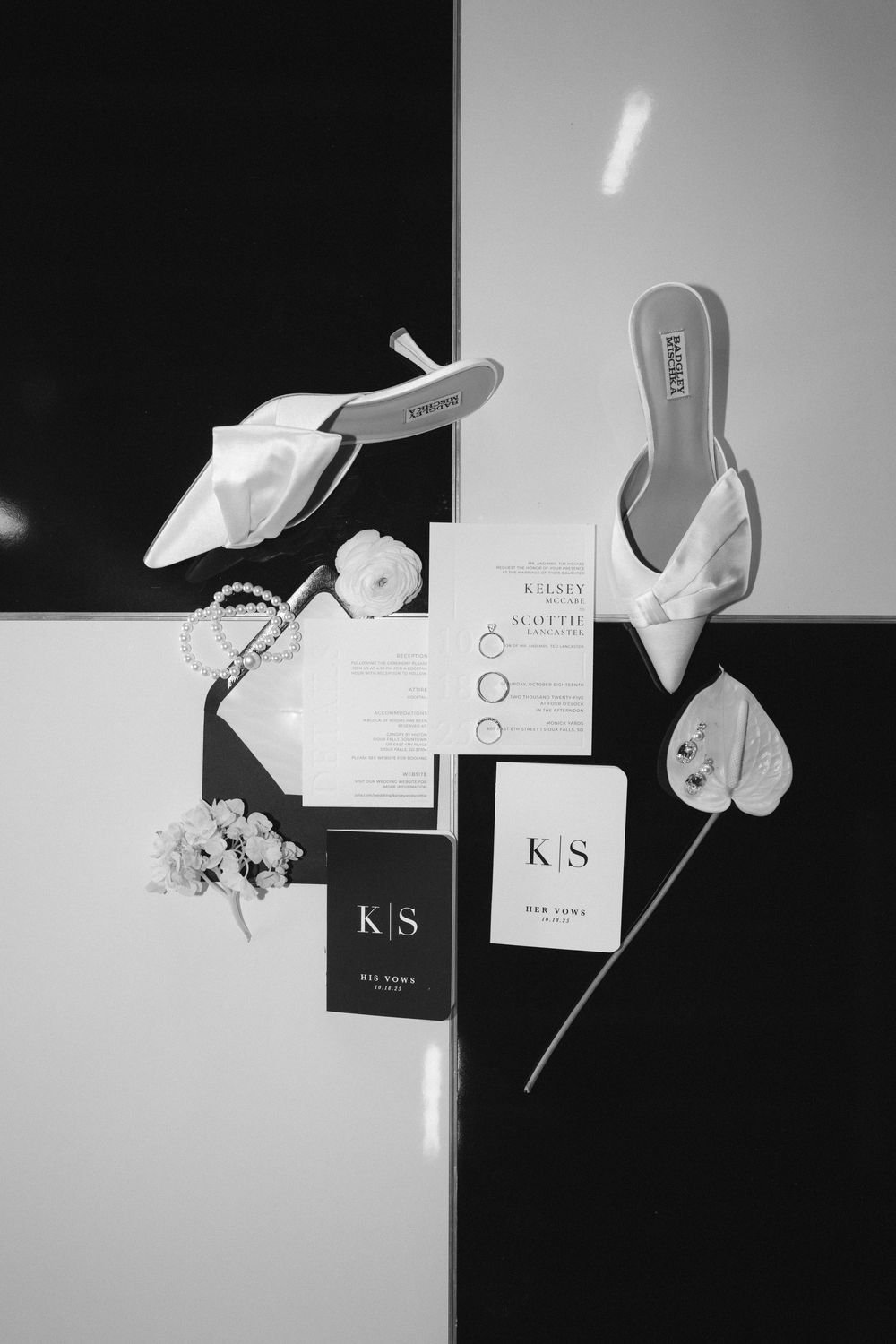 Black and white flat lay of wedding stationery, heels, rings, and floral details for a K&S wedding.