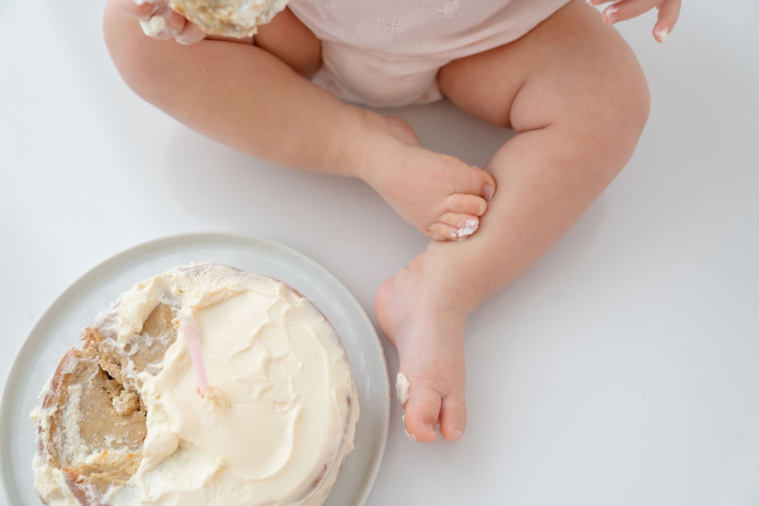 Baby sitting next to a smashed first birthday cake with white frosting on a white background.