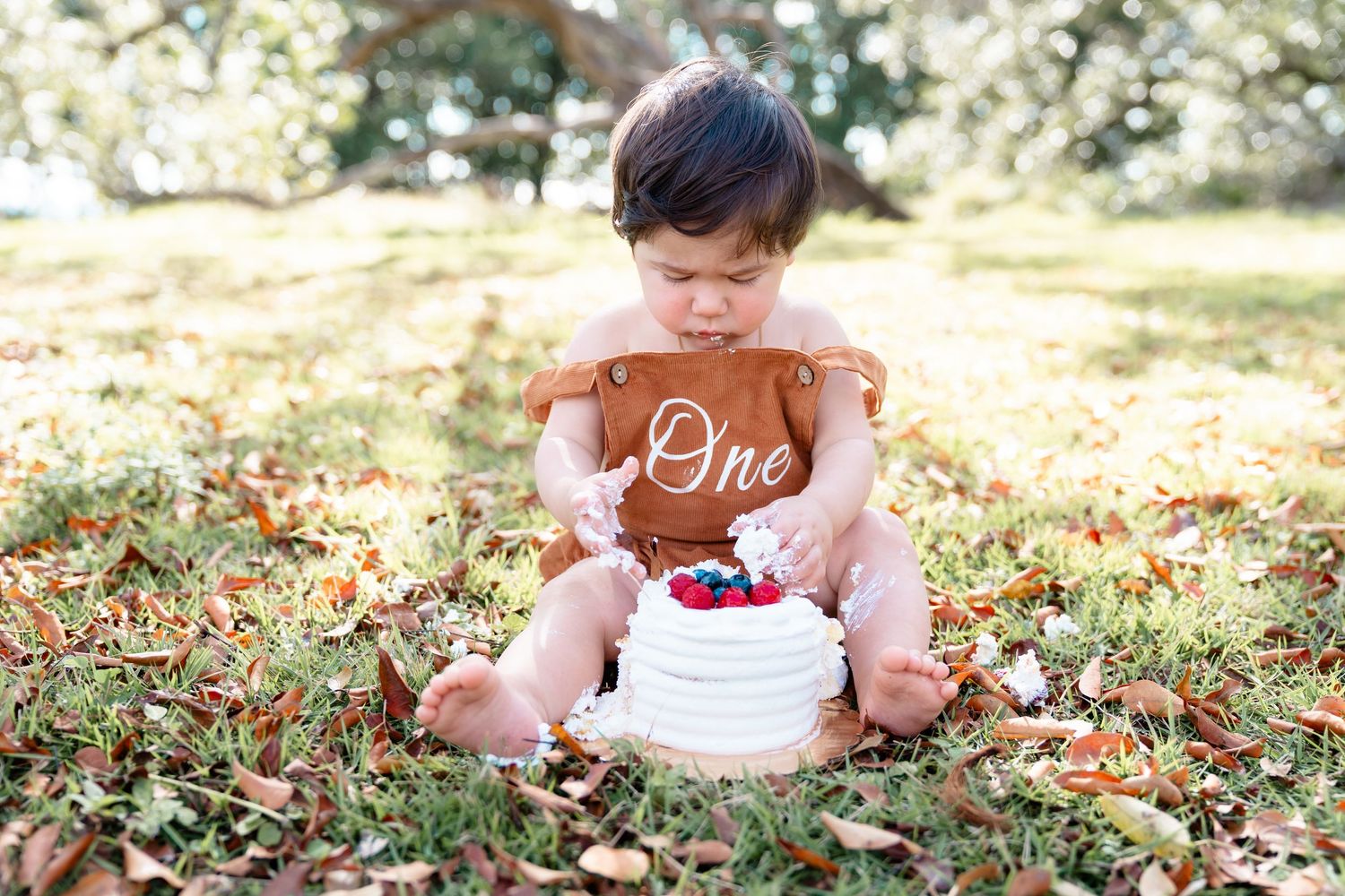 Baby girl in pink gingham dress and coral flower headband sitting on grass, eating cake with her hands.