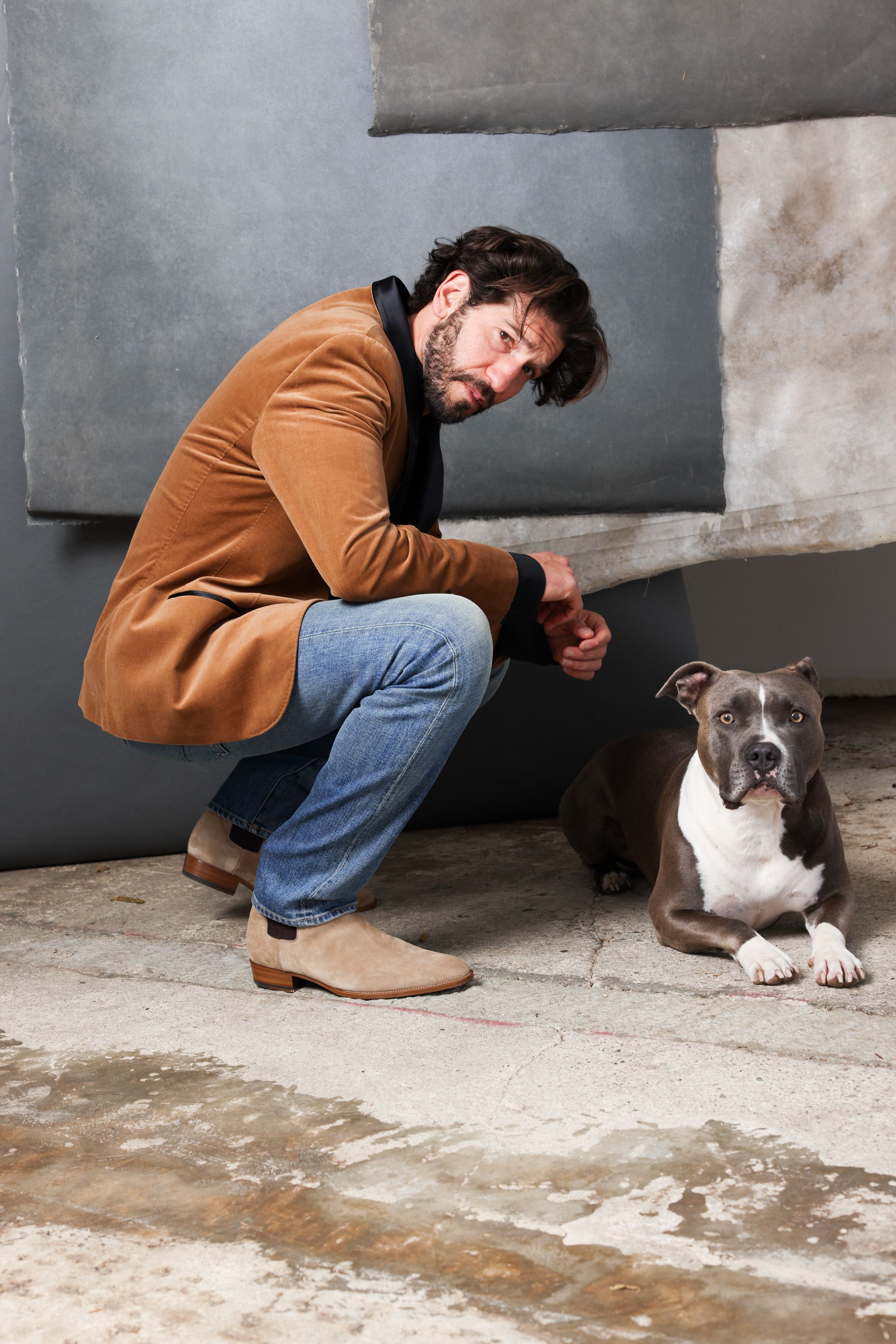 Actor Jon Bernthal in a brown jacket crouches down to interact with a gray and white pit bull dog in a concrete space.