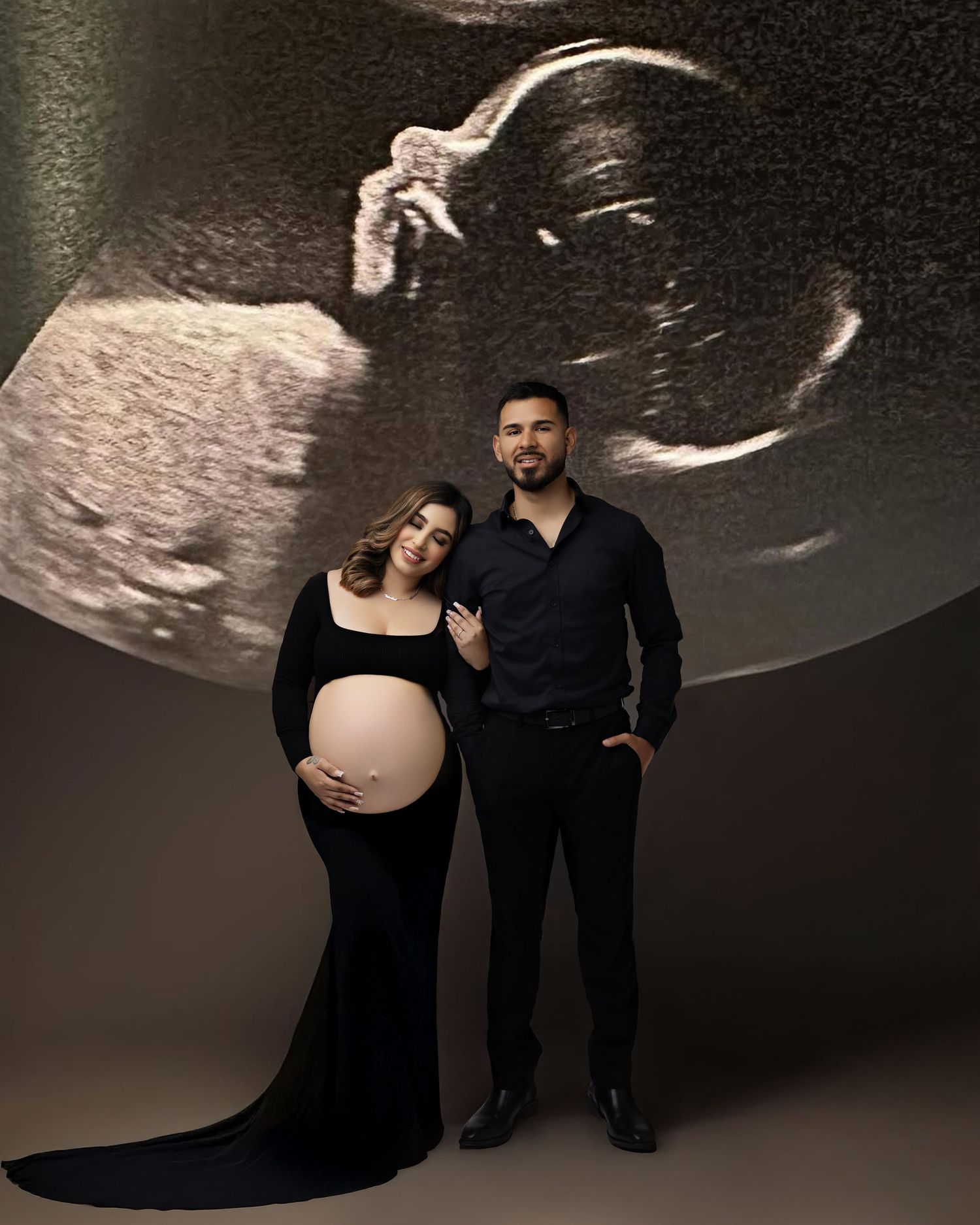 Pregnant couple in black outfits posing for maternity photo with ultrasound image projected behind them.