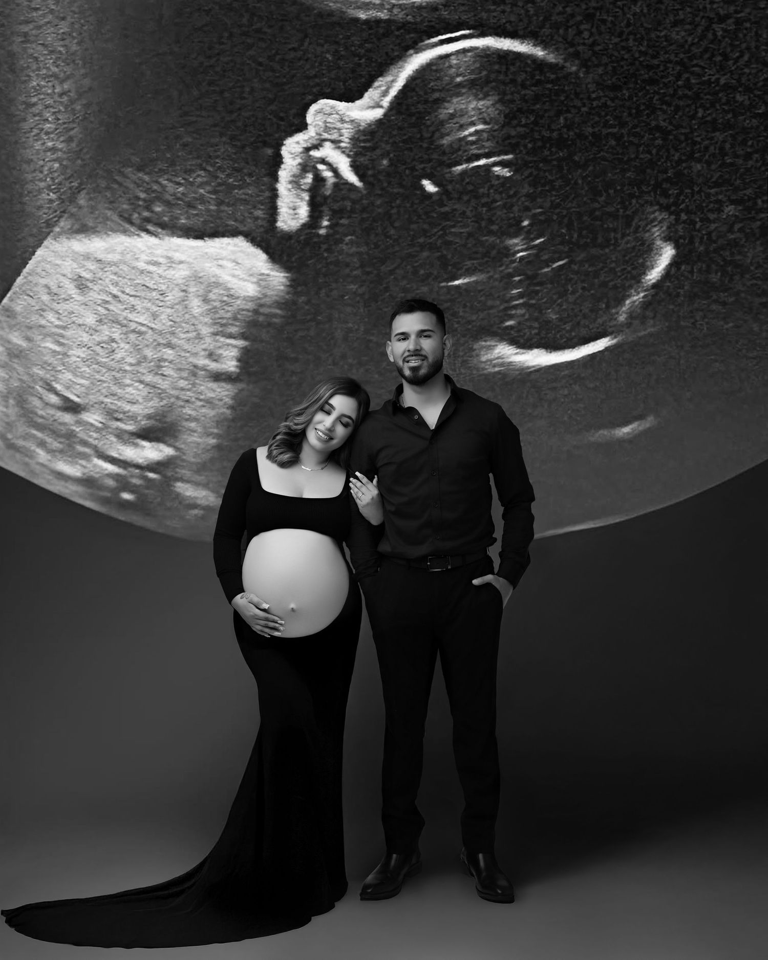 Black and white maternity photo of pregnant couple posing in front of projected ultrasound image in studio.