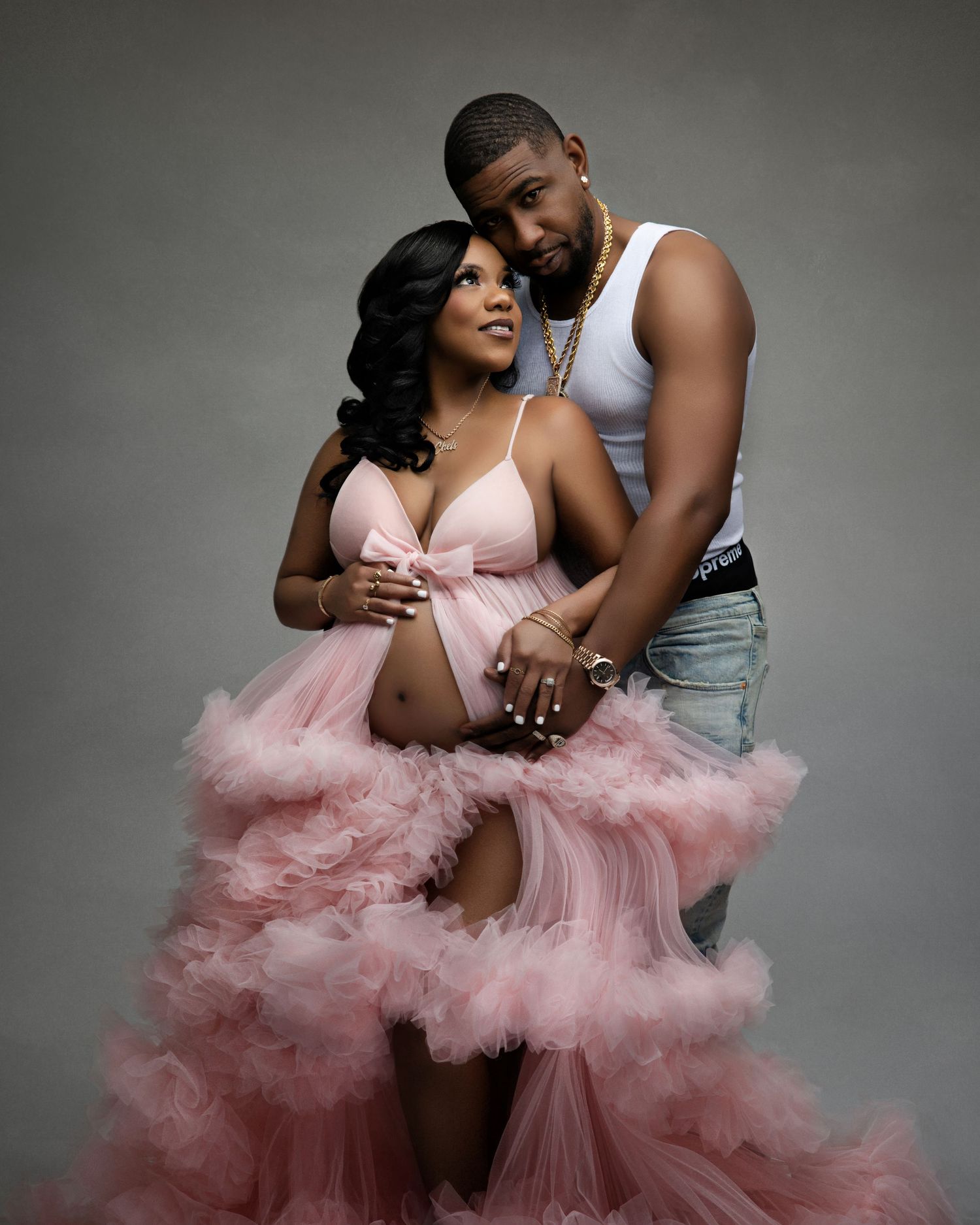 Couple poses for maternity photo shoot, woman in pink tulle gown cradling baby bump, man in white tank top embracing her.