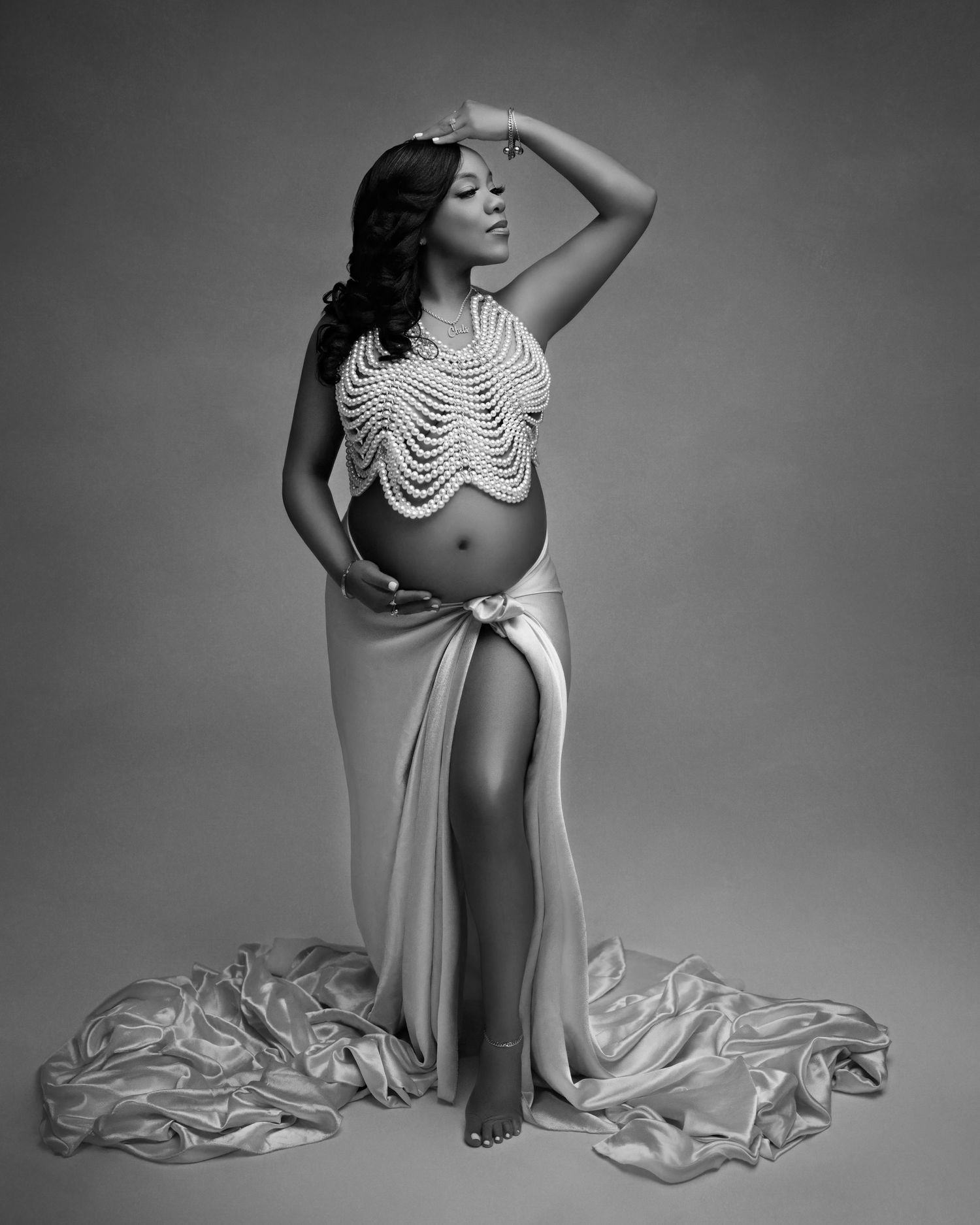 Black and white maternity photo of a pregnant woman in a pearl crop top and satin skirt with a thigh-high slit.