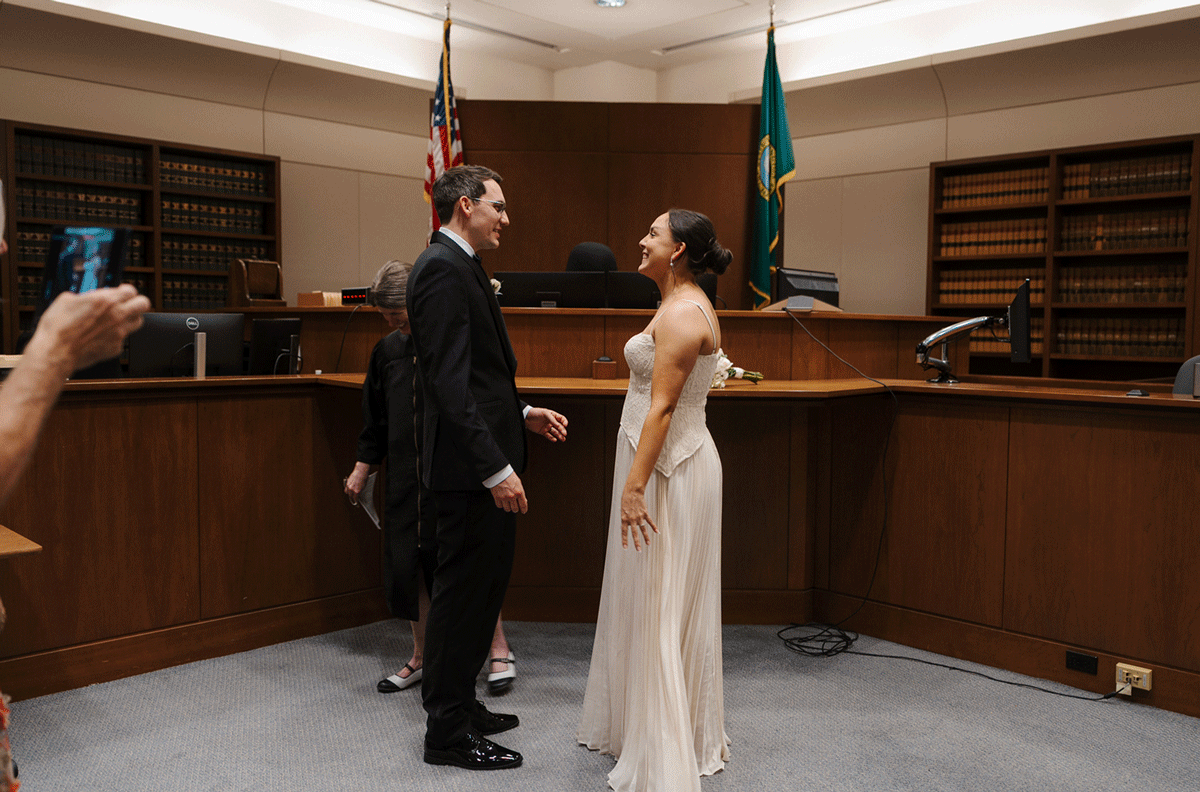 Couple exchanging vows in a courtroom civil wedding ceremony, man in black suit, woman in white gown.