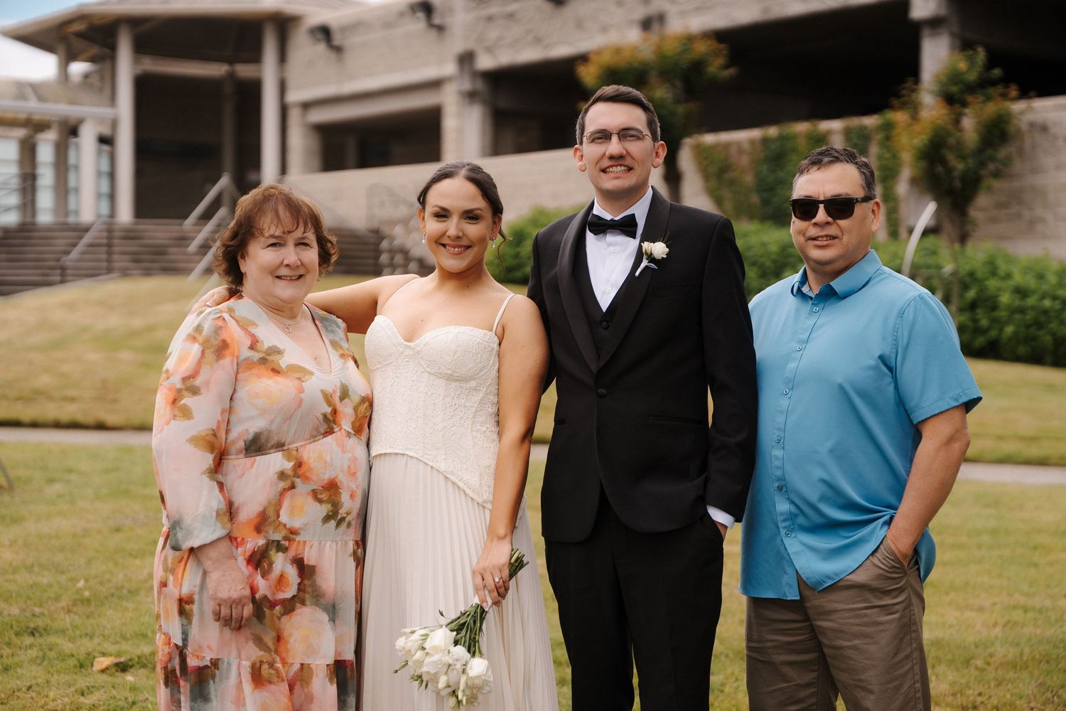 Newlywed couple in wedding attire poses outdoors with two family members in front of modern building.