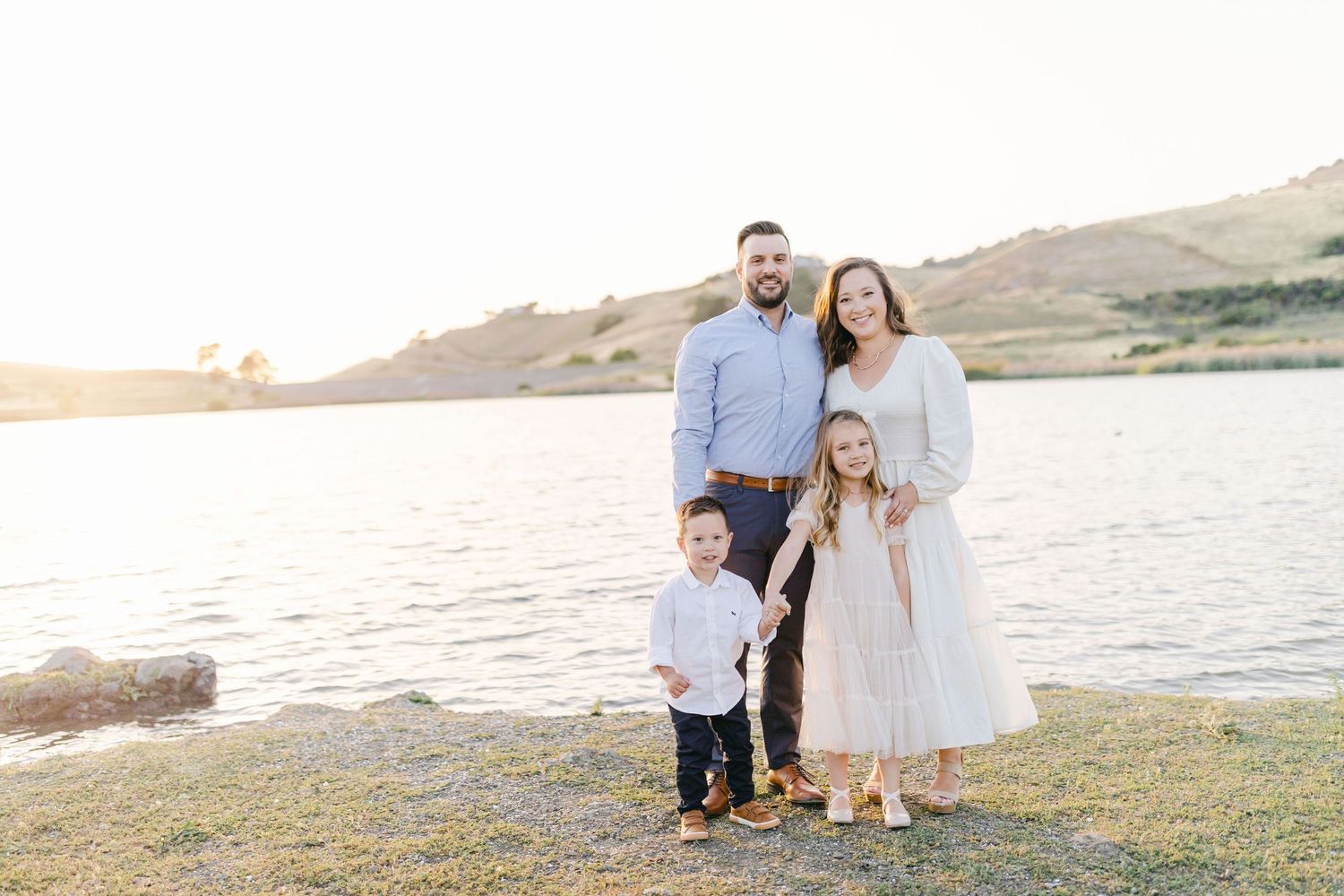 Family of four posing by a serene lake at golden hour, parents standing behind two young children on green grass.