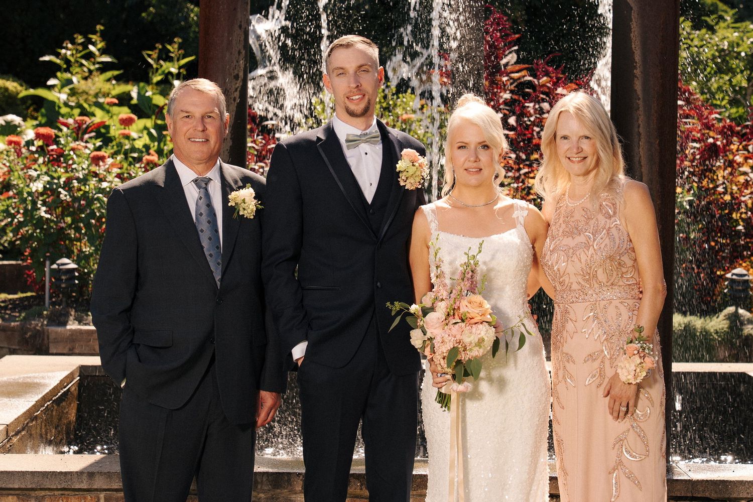 Wedding party of four posing in front of a fountain, bride holding bouquet in white gown with groom and family.