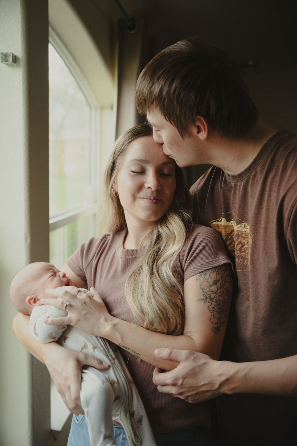 A young couple stands by a window, the man kissing the woman's head as she smiles and holds their newborn baby.