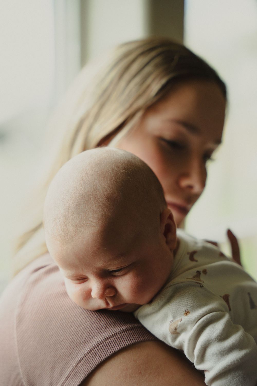 A young blonde mother tenderly holds her sleeping newborn baby against her shoulder near a window.