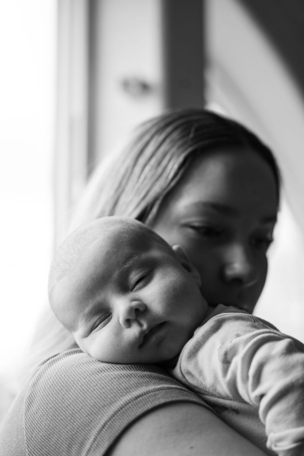 Black and white photo of a mother tenderly holding her sleeping newborn baby close to her chest near a window.