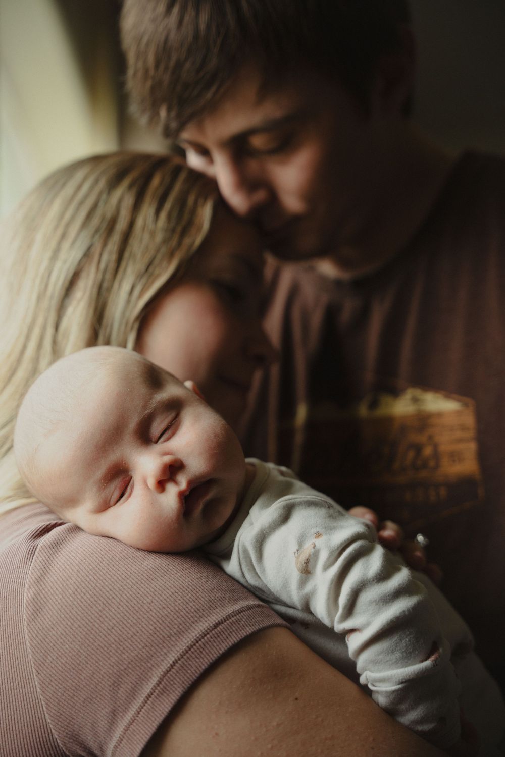 A newborn baby sleeps peacefully in mother's arms while father leans in to kiss the couple tenderly.