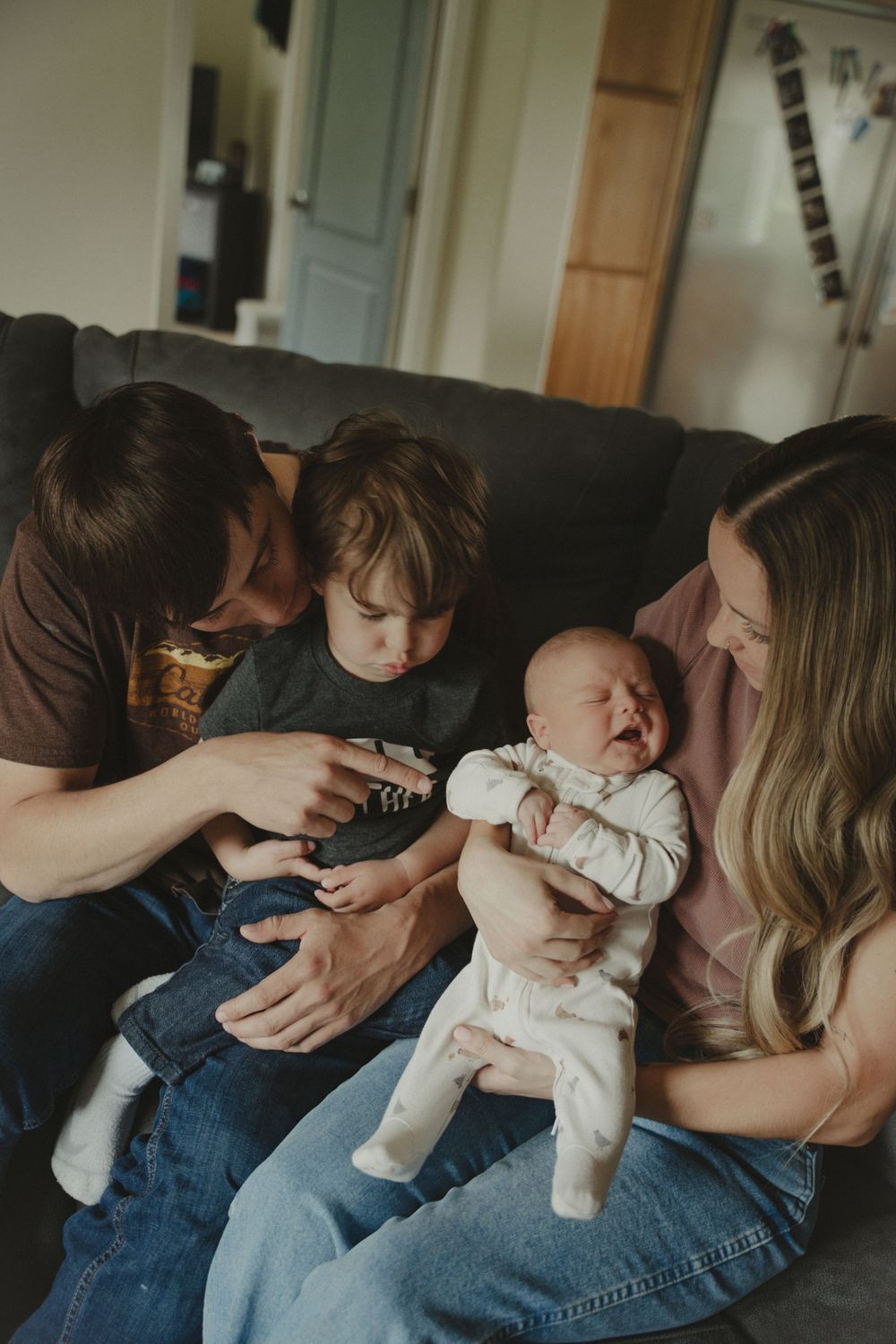 Family of four sitting on couch, parents smiling at crying newborn baby while young toddler looks on curiously.