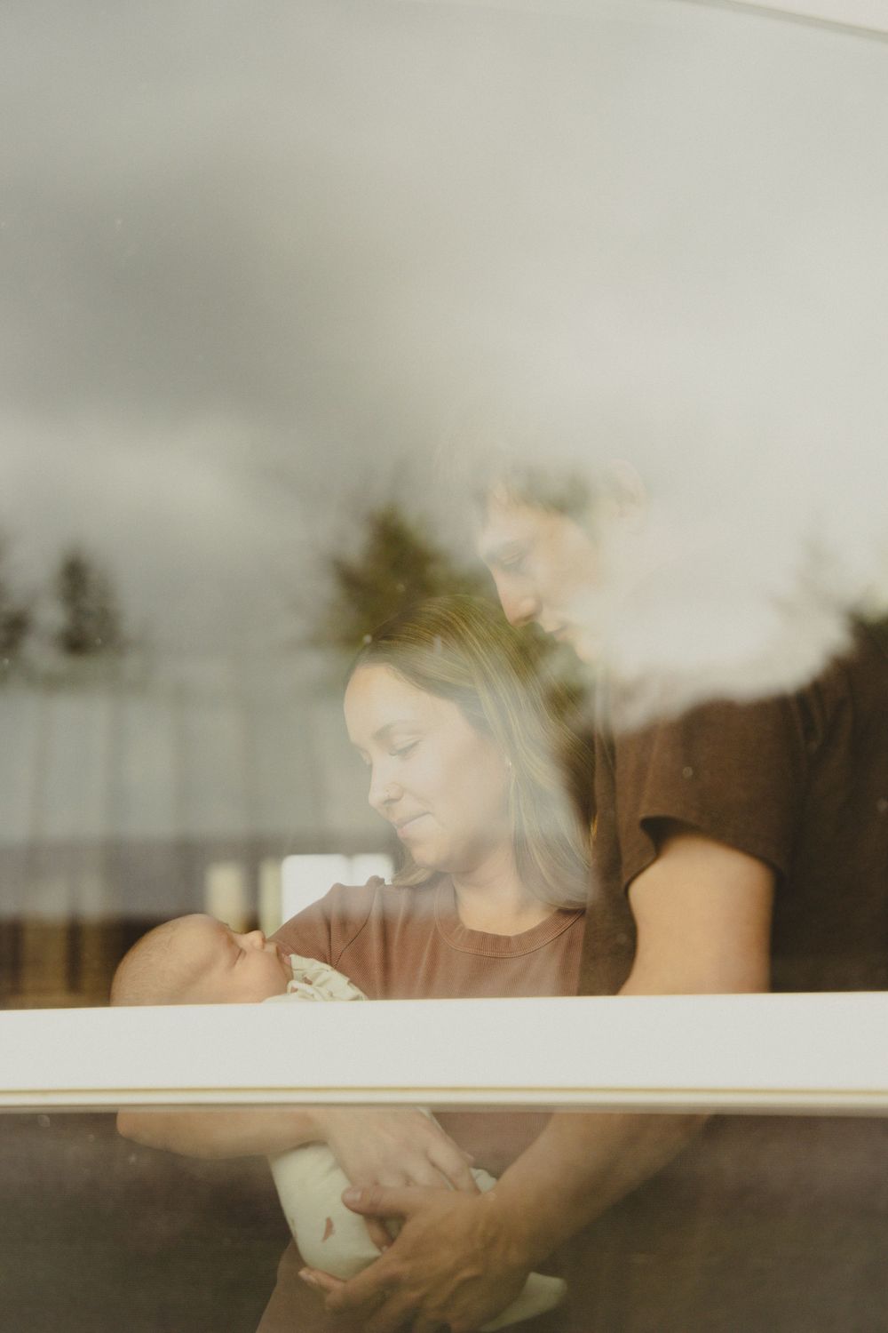 A couple tenderly gazes at their newborn baby, photographed through a window with soft natural light.