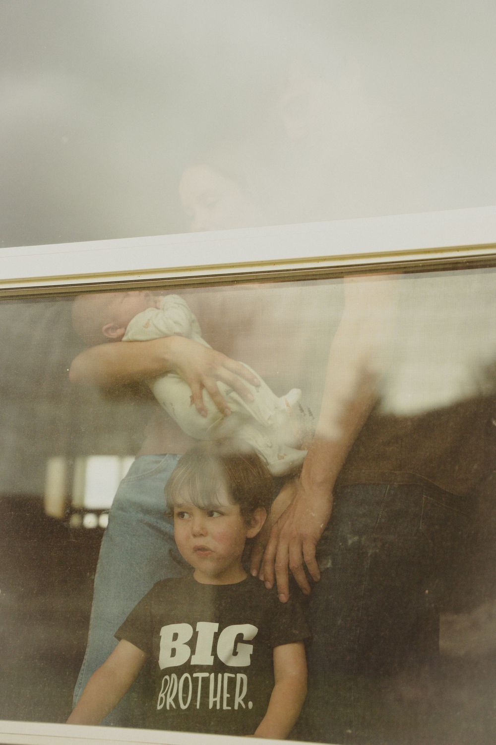 Young boy in Big Brother shirt stands before window with adult holding newborn baby behind him.