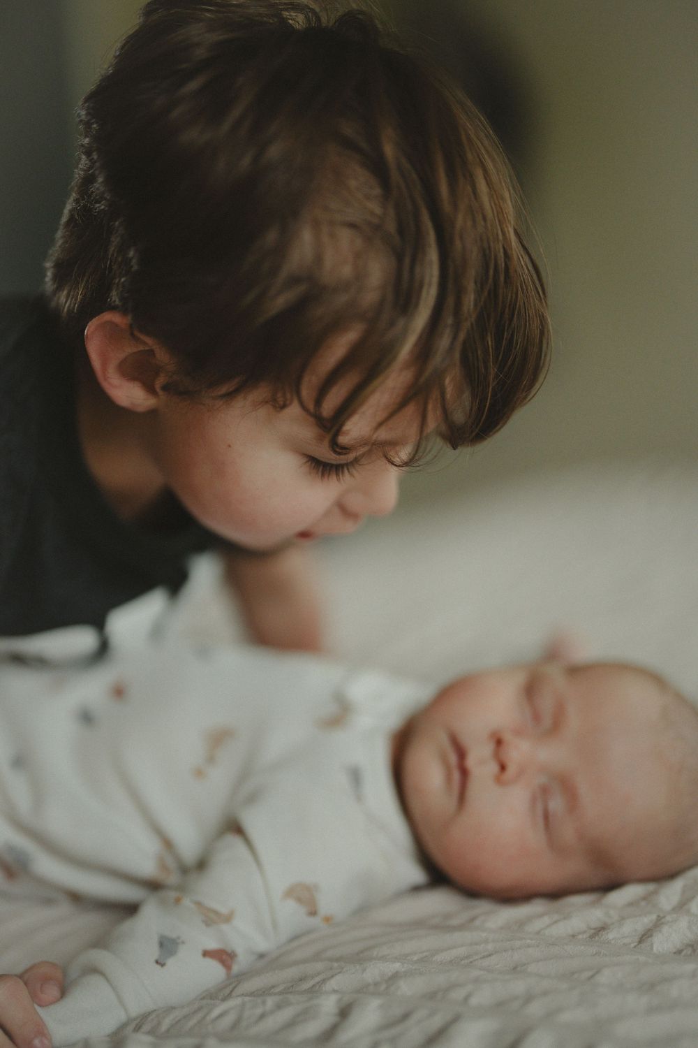 Young child gently kissing sleeping newborn baby sibling lying on white bedding.