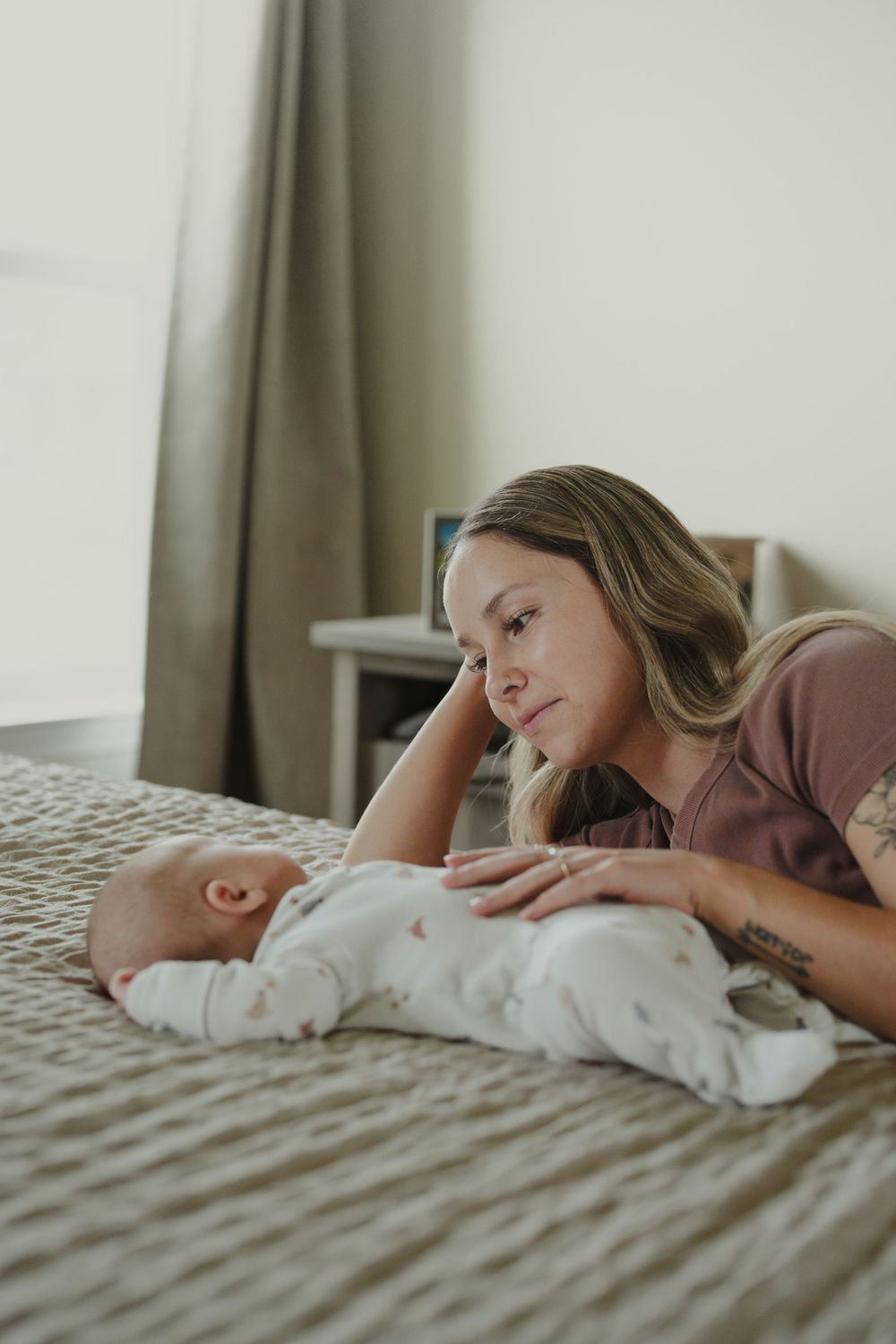 Mother lying on bed gazing lovingly at her newborn baby dressed in white onesie.