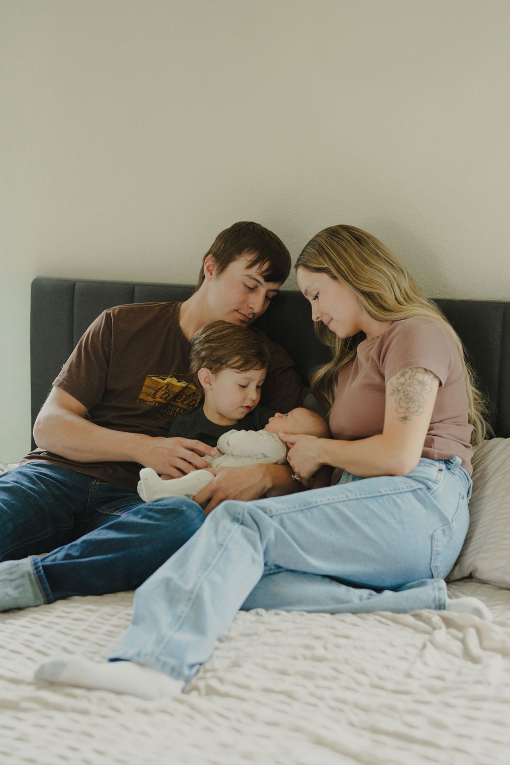 A young family sits together on a bed, parents lovingly gazing at their newborn baby held by the mother.