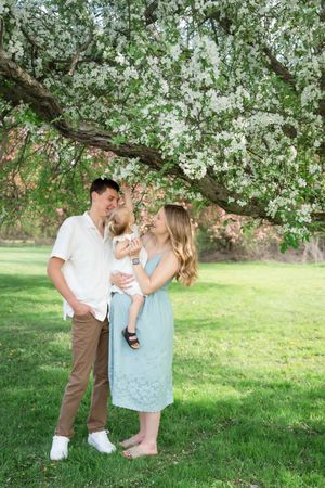 A young family with a baby stands under a blooming white flower tree on a green lawn in spring.