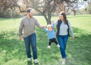 A happy family swings their toddler between them while walking in a lush green park with autumn trees.
