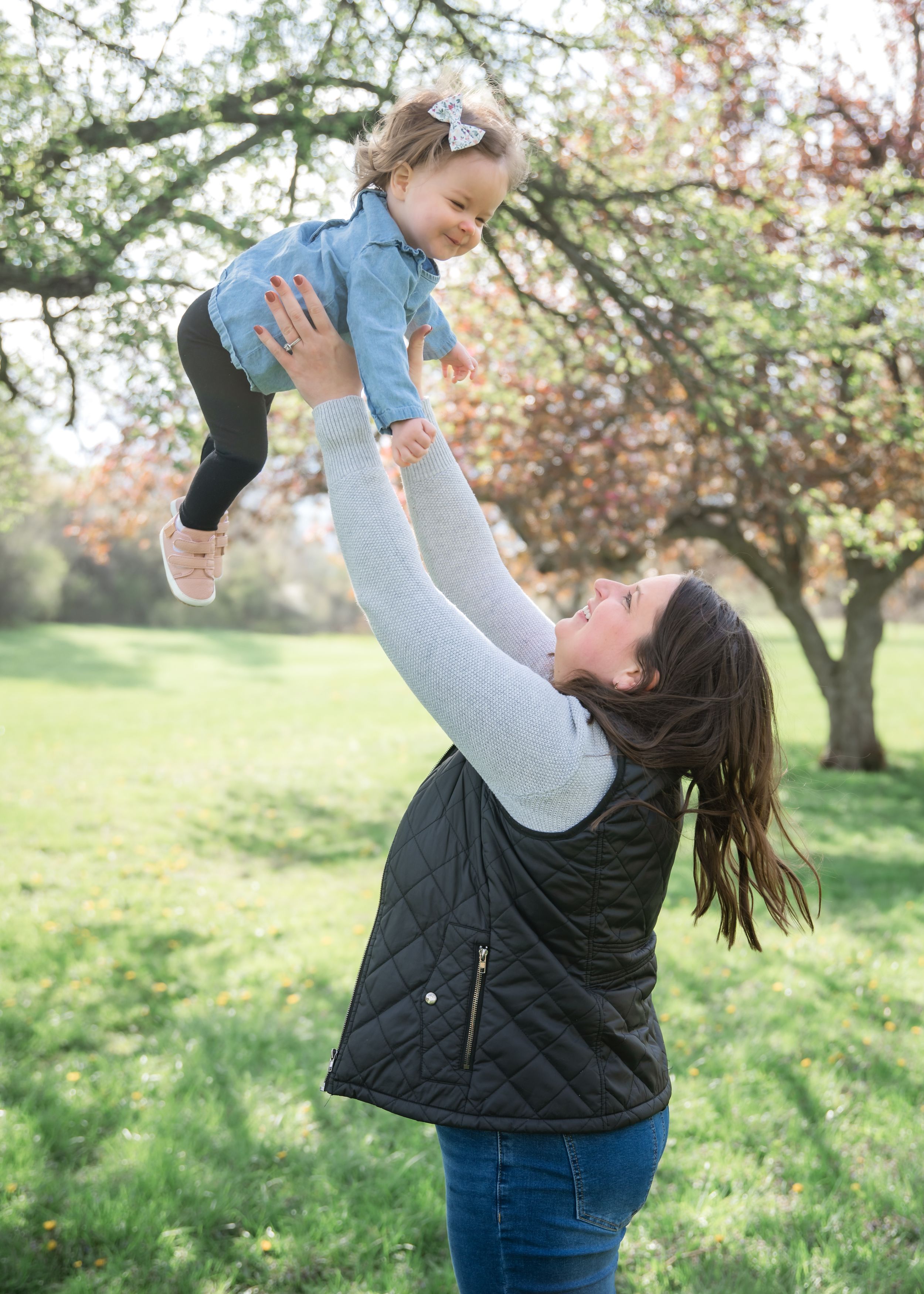A mother joyfully lifts her toddler daughter into the air outdoors among blooming spring trees.