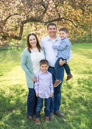 Family of four posing outdoors under blooming spring trees on green grass, parents with two young boys smiling.