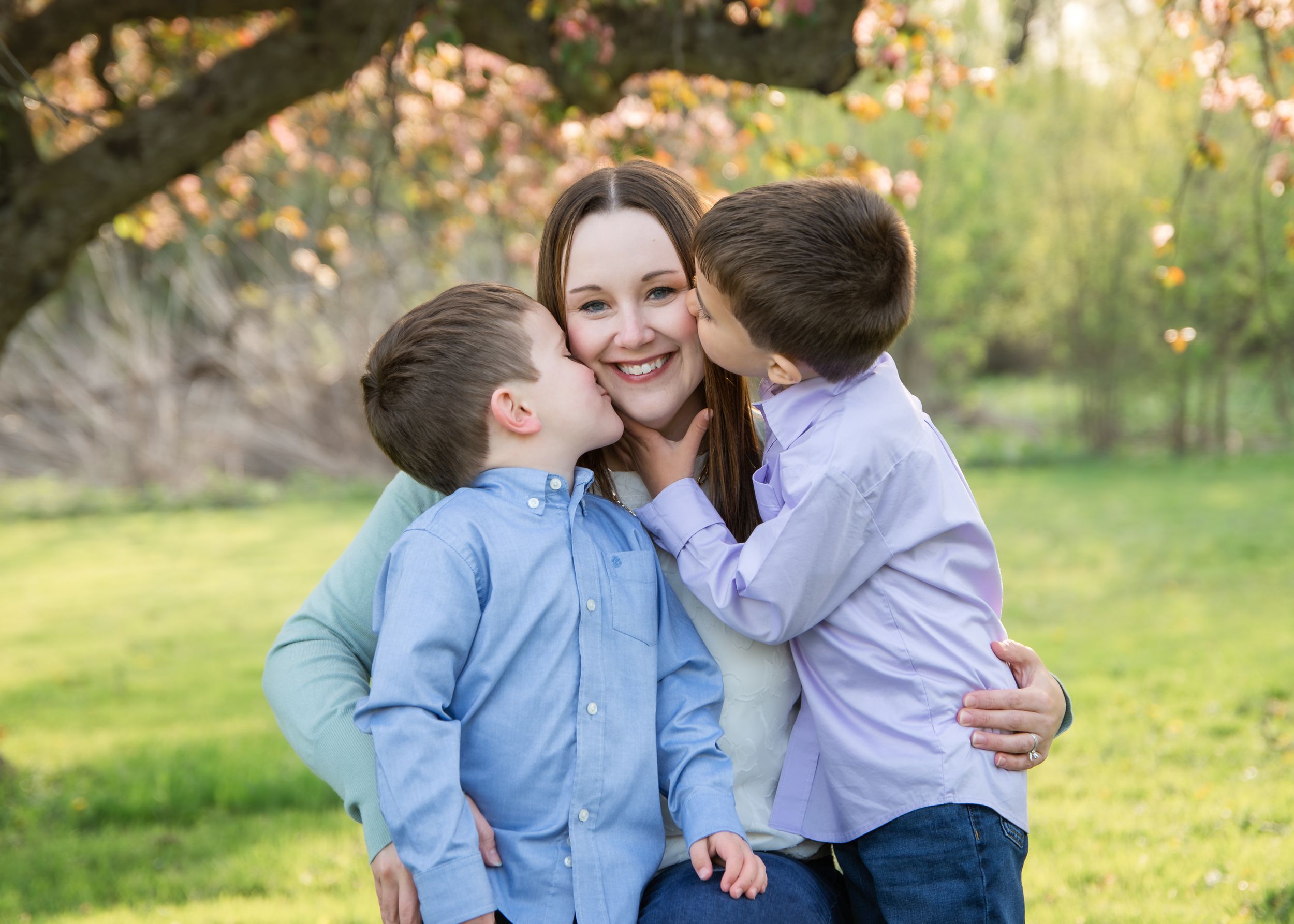 A smiling mother receives kisses on both cheeks from her two young sons outdoors under a blooming tree.