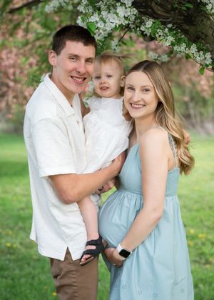 Young couple with toddler posing under blooming spring tree, woman in blue dress visibly pregnant, outdoor family portrait.