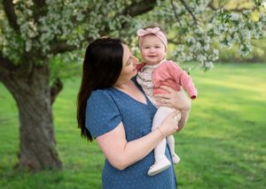 A mother holds her smiling baby girl under a blooming tree in a lush green park during spring.