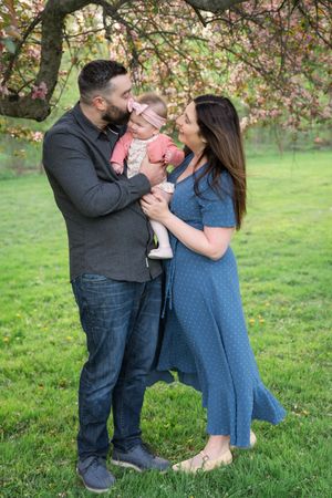 A happy family with a baby poses under blooming pink trees on green grass in spring.