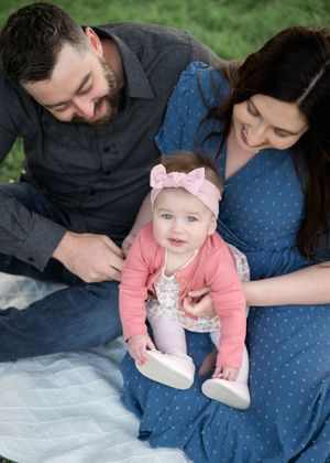 A happy family portrait with parents smiling at their baby girl in a pink outfit and bow headband on a blanket outdoors.