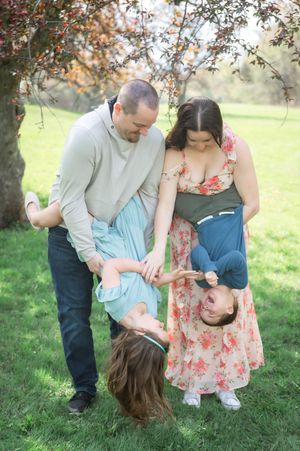 Family of four playing outdoors, parents playfully holding children upside down while laughing in a lush green park setting.