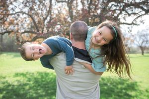 A father playfully holds two laughing children outdoors in a green park with blooming trees in the background.