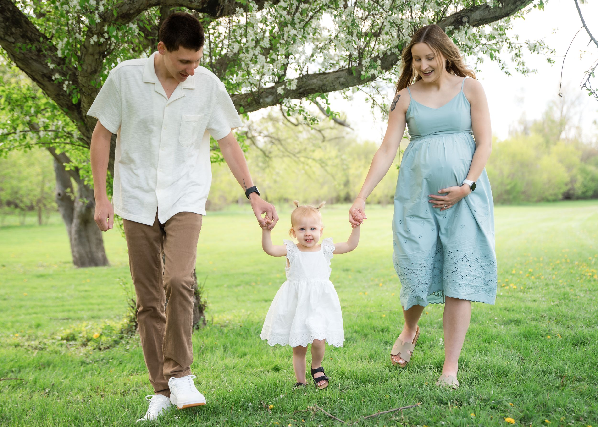 Pregnant woman and man hold toddler girl's hands walking in green park under blooming tree on sunny day.