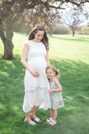 Pregnant woman in white dress with young daughter in floral dress standing on green grass outdoors.