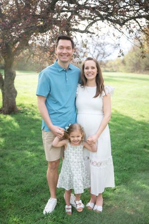 A happy family of three poses outdoors, with a pregnant woman in white, a man in blue, and a young girl in a floral dress.