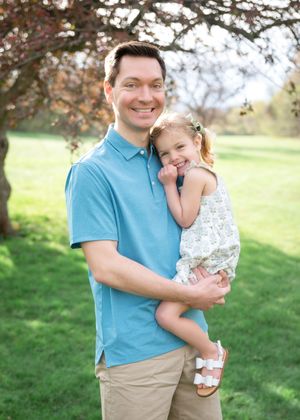 A smiling man in a blue polo shirt holds a happy young girl in a floral dress outdoors in a green park.